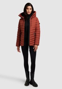 Puffed red-brown jacket with a hood, zip closure, and side pockets. Worn over a black top and leggings, completed with black boots.