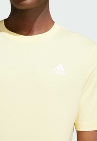 Camiseta de algodón amarillo claro con cuello redondo y mangas cortas, con un logo bordado de Adidas en blanco en la parte superior izquierda del pecho.