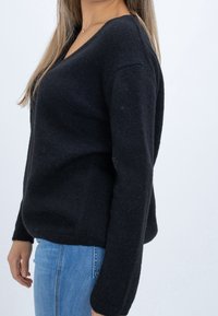 Schwarzer gestrickter Pullover mit V-Ausschnitt, strukturierter Stoff und gerippten Bündchen. Über einer hellblauen Jeans getragen, seitliche Ansicht.
