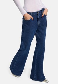 Jeans in denim a gamba svasata di colore blu scuro, con una texture liscia, tasche frontali, chiusura a bottone in metallo e leggeri dettagli di cucitura.