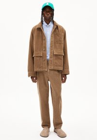 Brown corduroy jacket with two pockets, light blue button-up shirt, brown corduroy pants, and tan footwear. Green cap with logo detail.