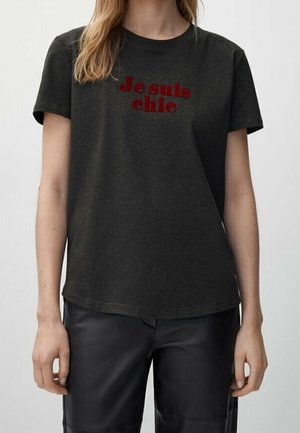 Personne portant un t-shirt gris foncé avec un texte rouge "Je suis chic" et un pantalon noir, debout devant un fond clair uni.