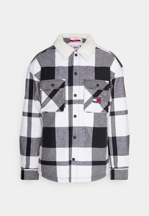 Hombre con chaqueta a cuadros en blanco y negro con cuello de borrego, camisa azul claro, pantalones blancos y zapatillas azules claras de pie.