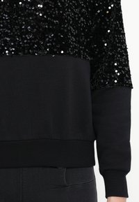 Sweatshirt preto com uma parte superior de lantejoulas e uma parte inferior lisa. Mangas longas com punhos canelados, design minimalista.