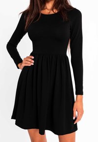 Femme portant une robe noire ajustée à manches longues avec une jupe froncée, main sur la hanche, et un collier avec une croix visible au niveau du décolleté.