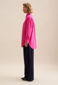Roze gestreepte blouse met hoge kraag, lange mouwen en knoopmanchetten, gecombineerd met marineblauwe brede broek en puntige zwarte schoenen.