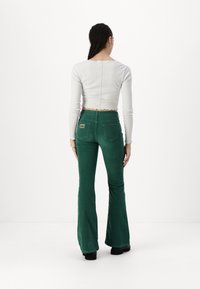 Green corduroy flared pants with back pockets and a branded label. The model wears a long-sleeve white top with a cropped, scalloped hem.