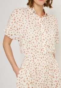 Kurzärmeliges Kleid in hellem Beige mit einem rosa gepunkteten Muster, versehen mit einem Knopfkragen und einem elastischen Taillenbund mit seitlichen Taschen.