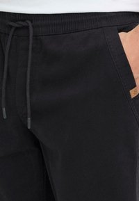 Joggers en coton noir avec une taille élastique, un cordon de serrage, des poches latérales et une petite étiquette de logo beige sur le côté. Texture douce.