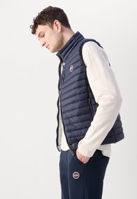 Vesta din material matlasat de culoare bleumarin, cu guler înalt și închidere cu fermoar, având un logo circular pe piept și buzunare laterale, purtată peste un pulover ușor.