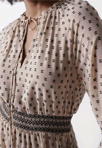 Beige mekko, jossa on mustia geometrisia kuvioita, rypistetty pääntie, silmukkapitsi kauluksessa ja smokkaukset vyötäröllä.