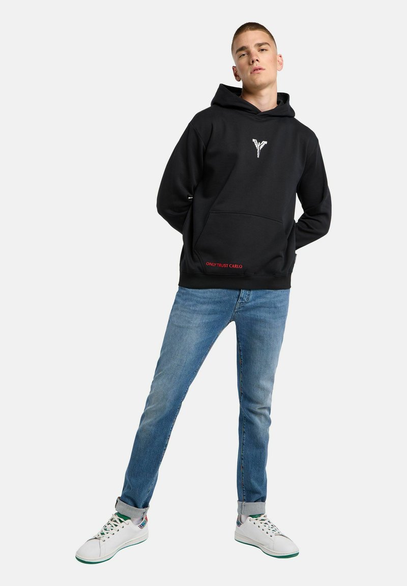 Schwarzer Kapuzenpullover mit einer Fronttasche, versehen mit einem weißen Grafikdesign auf der Brust und rotem Text nahe dem Saum. Getragen mit blauen Jeans und weißen Turnschuhen.