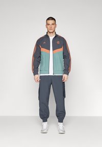 Giacca da palestra con pannelli grigio scuro, blu teal e arancione, con tre strisce sulle maniche. Abbinata a pantaloni cargo scuri e sneakers bianche.