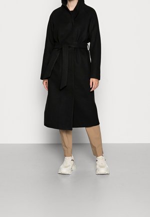 Person iført en lang sort bæltefrakke, beige bukser og hvide chunky sneakers, stående mod en ensfarvet lys grå baggrund.