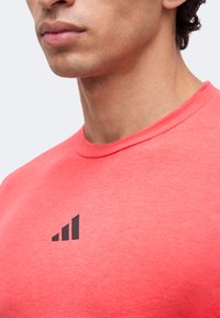 Gros plan d'une personne portant une chemise rouge corail avec un petit logo de graphique à barres noir sur la poitrine, montrant le cou et le bas du visage.