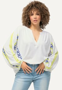 Hvid, løs siddende bluse med V-hals. Har blå og gule broderede mønstre på de lange ærmer. Båret med lyseblå jeans.