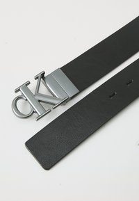 Cinto de couro preto com uma fivela metálica prateada com o logótipo "CK". O cinto possui furos perfurados para ajuste ao longo do seu comprimento.