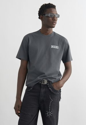 Hombre con camiseta "Dickies" gris oscuro, vaqueros negros con tachuelas, gafas de sol y cinturón negro, de pie con una mano en el bolsillo.