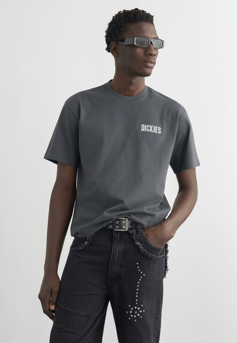 Hombre con camiseta "Dickies" gris oscuro, vaqueros negros con tachuelas, gafas de sol y cinturón negro, de pie con una mano en el bolsillo.