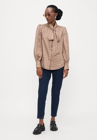 Bruine blouse met patroon, een strik om de hals, pofmouwen en knoopsluiting, gecombineerd met donkerblauwe jeans en zwarte instappers.