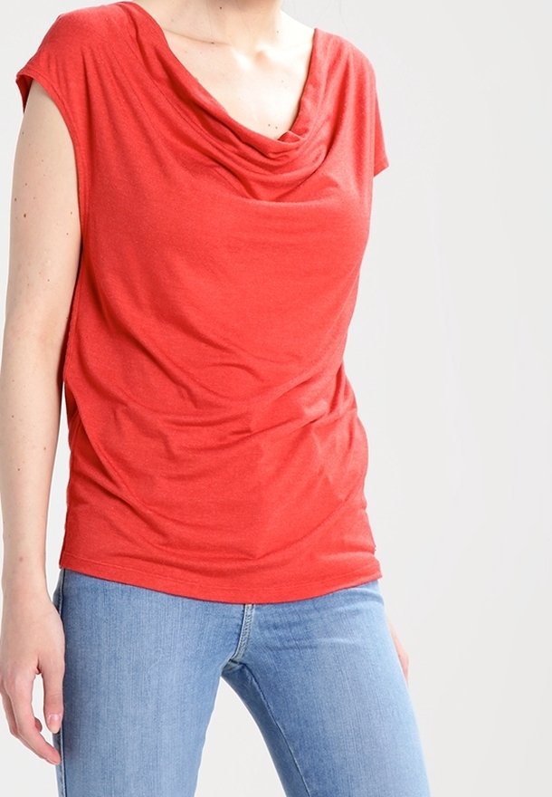 Personne portant un haut rouge ample à manches courtes avec un col drapé et un jean bleu clair sur un fond blanc uni.
