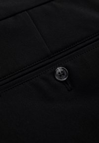 Musta kangas, jossa on sileä pinta, sisältää käytännöllisen nappidetailin. Kaksi vaakasuoraa taskua on näkyvissä, esitellen yksinkertaisia muotoiluelementtejä.