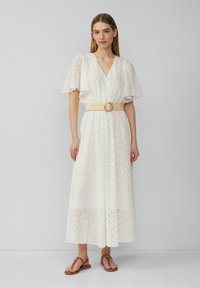 Weißes Maxikleid mit kurzen, ausgestellten Ärmeln, V-Ausschnitt und einem strukturierten Muster. Inklusive einem breiten beigen Gürtel mit einer runden Schnalle. Braune Sandalen.