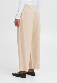 Pantaloni beige a gamba larga con vita elasticizzata, due tasche posteriori e una texture liscia, abbinati a scarpe nere flat.