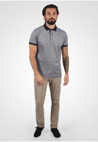 Šedé polo tričko s černým límečkem a rukávy, doplněné béžovými kalhotami a černými teniskami. Jednoduchý design, krátké rukávy a knoflíky.