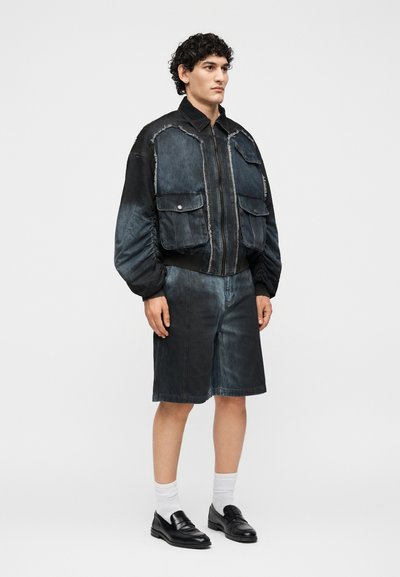 Νέος άνδρας με σκούρο πλυμένο bomber τζιν μπουφάν με μεγάλες μπροστινές τσέπες και ταιριαστά σορτς μέχρι το γόνατο, μαύρα μοκασίνια και λευκές κάλτσες.