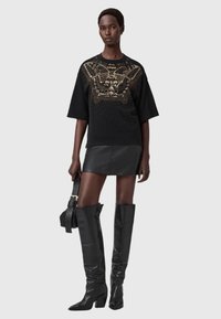 Sudadera negra con diseño gráfico dorado y adornos, combinada con una minifalda de cuero negra y botas negras de tacón hasta la rodilla.