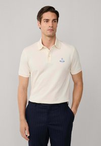 Cremfarbener Poloshirt mit kurzen Ärmeln, Knopfleiste mit drei Knöpfen und aufgesticktem blauen Logo auf der Brust, kombiniert mit dunkelblauen gestreiften Hosen.