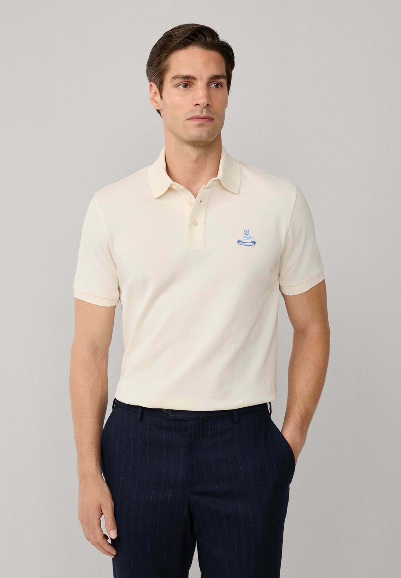Cremfarbener Poloshirt mit kurzen Ärmeln, Knopfleiste mit drei Knöpfen und aufgesticktem blauen Logo auf der Brust, kombiniert mit dunkelblauen gestreiften Hosen.