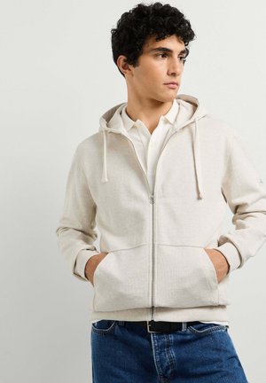 Junger Mann mit lockigem Haar, der einen hellbeigen Zip-up-Hoodie über einem weißen Hemd mit Kragen und blauen Jeans trägt, die Hände in den Taschen des Hoodies, schaut seitlich.