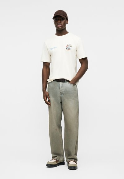 Νεαρός άνδρας που φοράει κρεμ κοντομάνικη μπλούζα με σχέδια, χαλαρά ξεβαμμένα γκρι τζιν, καφέ καπέλο και μπεζ παπούτσια με κορδόνια και μαύρες σόλες.