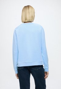 Personne aux cheveux blonds courts portant un sweat-shirt bleu clair et un jean foncé, debout de dos devant un fond blanc uni.