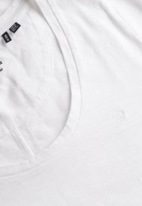 T-shirt en coton blanc à col rond, coutures visibles, étiquette de taille (UK 10, US 6) et logo brodé subtil sur la poitrine.