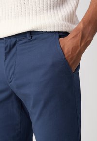 Pantalon en coton bleu marine avec une coupe droite. Il présente une poche latérale et une texture lisse, associé à un pull en tricot côtelé crème au-dessus.