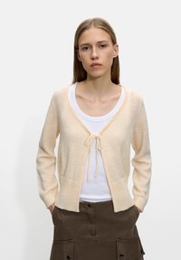 Jasnokremowy kardigan z zapięciem na wiązanie, z prążkowanymi mankietami i dołem, noszony na białej koszuli. Miękka faktura i lekka, dziana konstrukcja.