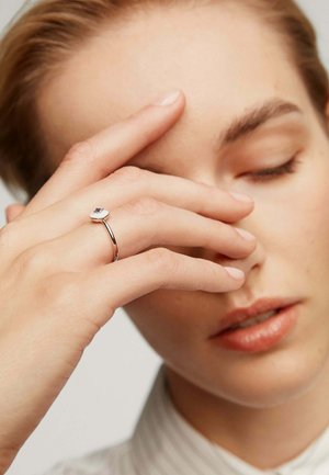 Anillo de plata con una banda lisa y una piedra preciosa redonda de color claro. La mano del modelo cubre parcialmente su rostro, enfatizando el anillo.