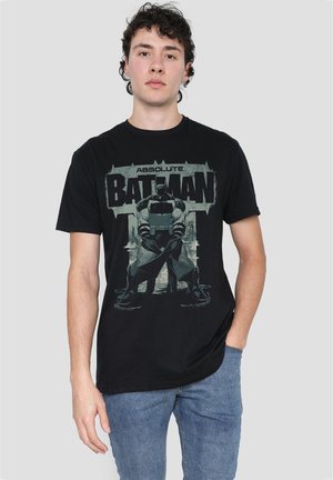 Young man wearing a black Batman graphic t-shirt and blue jeans, standing against a plain light gray background.