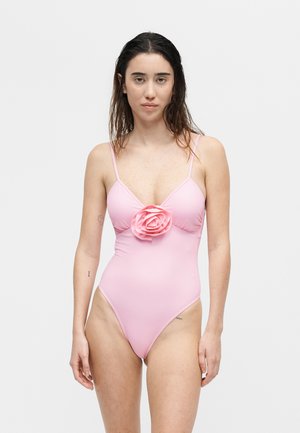 Γυναίκα με βρεγμένα μαλλιά που φοράει ανοιχτό ροζ ολόσωμο μαγιό με λεπτές τιράντες και μεγάλο λουλούδι τριαντάφυλλου στο κέντρο μπροστά.