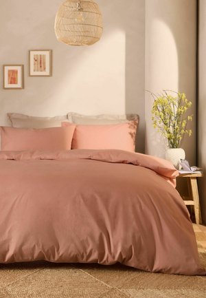 Letto matrimoniale con biancheria e cuscini color pesca, parete beige con due opere d'arte incorniciate, lampada a sospensione intrecciata, tavolino in legno con vaso di fiori gialli.