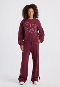 Bordeaux collegepaita ja housut, joissa on suuri "89" printti, rento istuvuus ja kontrastiväriset valkoiset sivunauhat housuissa. Pehmeä kankaan rakenne.