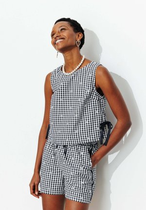 Femme souriante regardant vers le haut, portant un haut sans manches à carreaux noir et blanc et un short assorti avec un collier de perles et des boucles d’oreilles en forme de cœur.