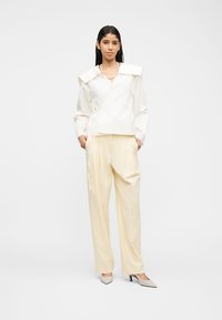 Γυναίκα που φοράει ανοιχτόλευκο τοπ τύπου wrap με φαρδιά γιακά, εκρού παντελόνι με φαρδιά πόδια, μπεζ τακούνια και μαργαριταρένιο κολιέ, στέκεται με τα χέρια στις τσέπες.