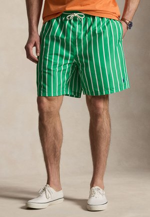 Mann iført grønne badeshorts med hvite vertikale striper, kombinert med hvite joggesko og en oransje skjorte, stående med hendene i lommene.