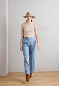 Beige ärmlös topp, högmidjade ljusblå jeans och bruna anklarstövlar. Modellen bär en stråhatt. Slät vägg och trägolv.