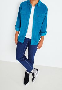 Camicia button-up teal con due tasche sul petto, abbinata a jeans in denim scuro con cuciture a contrasto e sneakers in bianco e nero.
