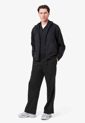 Giacca leggera nera con cappuccio, abbinata a una camicia nera con bottoni e pantaloni neri larghi. Sneakers grigie con dettagli in rete.